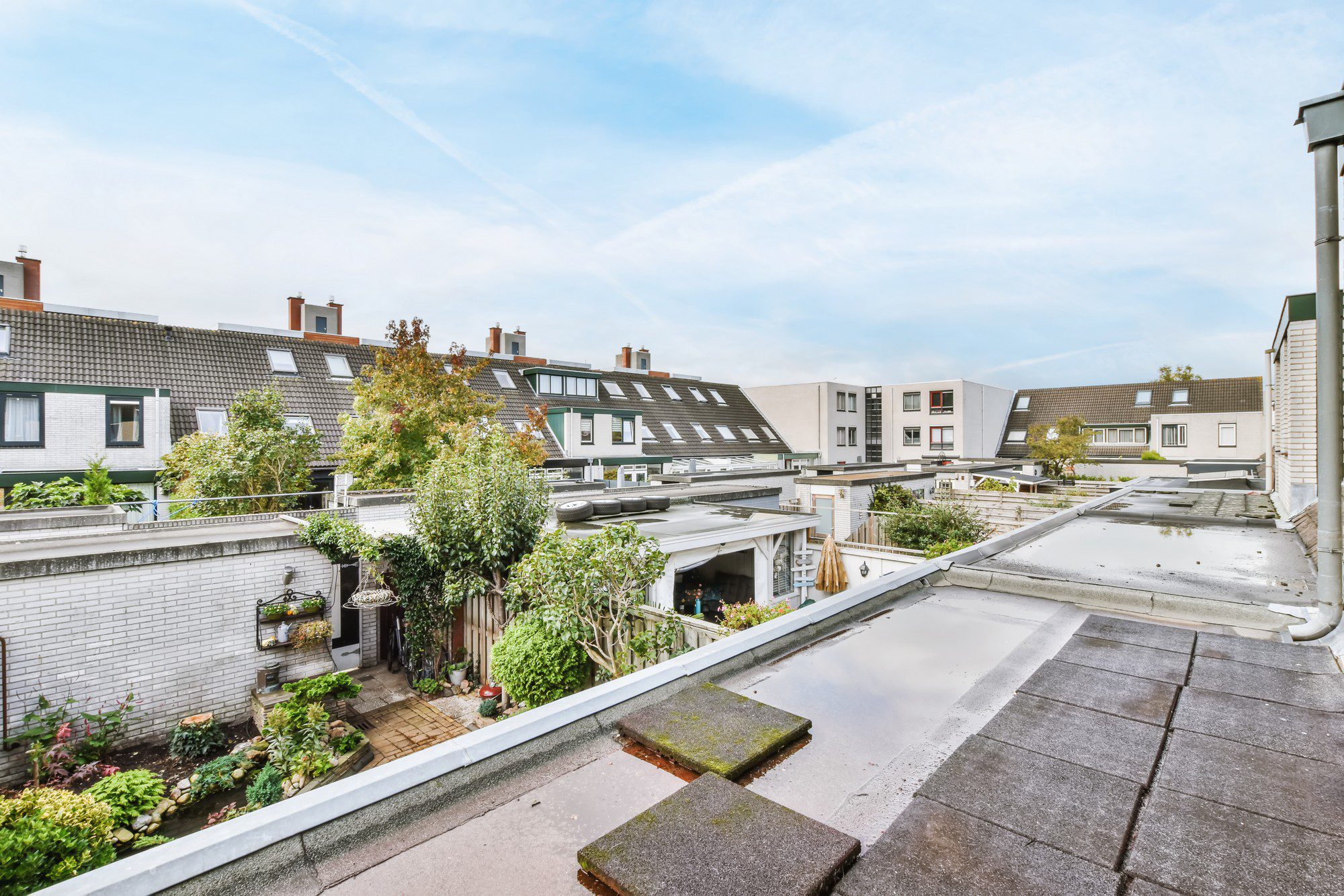 The image shows a residential area with townhouses or similar connected properties. In the foreground, there is a flat rooftop possibly belonging to a garage or an extension of a house. This rooftop has paving stones and a water puddle, suggesting recent rain. In the mid-ground, there are backyards with various plants, trees, and shrubbery, indicating that the homeowners have put effort into creating garden spaces. Visible in the backyards are also fences and garden decorations. Further back, the houses have pitched roofs with dormer windows and solar panels, highlighting a focus on sustainable energy. The sky is partly cloudy, suggesting a day with a mix of sun and clouds.