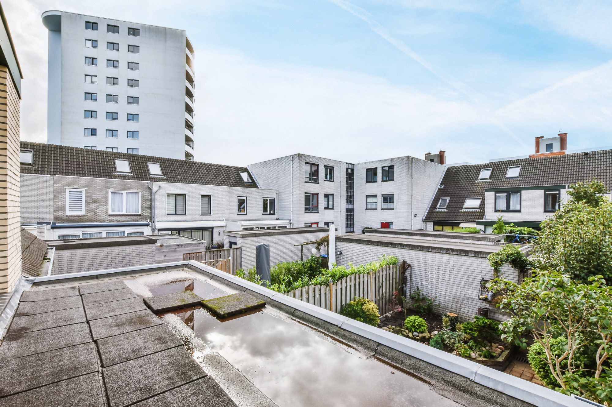 The image shows a view from a higher vantage point cascading down to a residential area. In the foreground, there is a flat rooftop with some standing water and a few slabs looking like pavers, indicative of a potential roof terrace area. Moving beyond, there are multiple residential buildings featuring gabled and flat roofs.

The architecture indicates an urban environment with closely spaced houses, some with dormer windows indicating attic or loft spaces. Between the buildings, there is a fenced-off area with lush greenery, likely a small private garden or communal green space showing well-maintained bushes and plants. In the distance, a tall white building with many windows looms over the smaller structures, contrasting with the lower-rise buildings in the foreground. The sky is partially cloudy, suggesting a typical overcast day. Overall, the scene portrays a dense urban residential neighborhood with mixed types of housing.