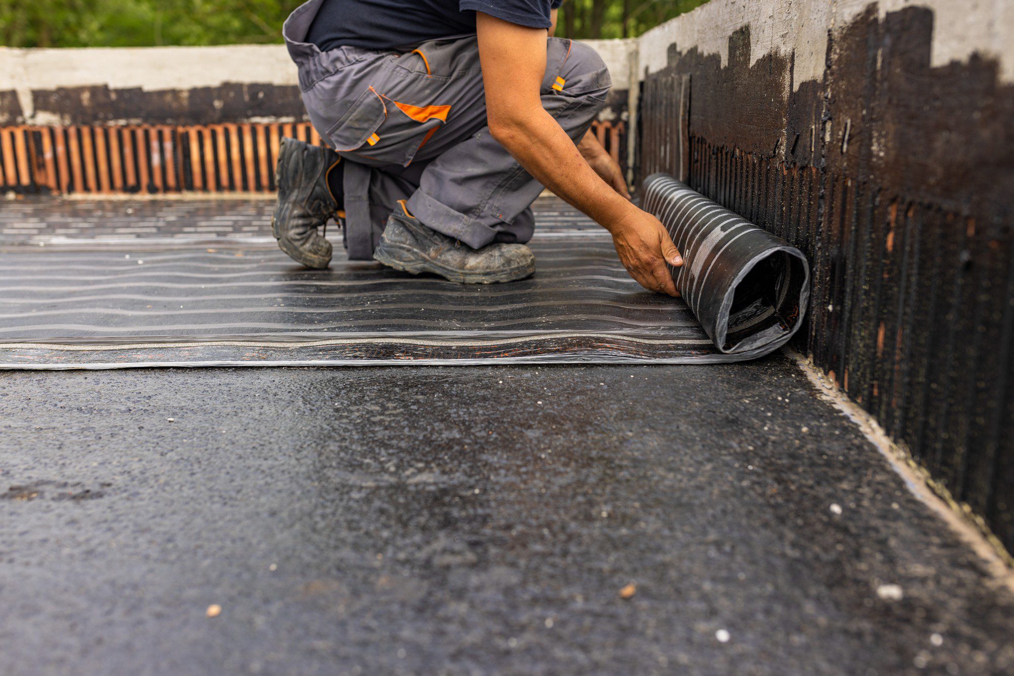 The image depicts a person in work clothes installing a waterproofing membrane on a flat surface, which appears to be part of a construction or renovation project. The individual is kneeling and appears to be unrolling or laying down the black corrugated material, which is likely used to prevent water ingress in the construction area. The person is wearing sturdy work boots and the work environment looks to be outdoors, as indicated by the daylight and the presence of vegetation in the background. The membrane is being applied over a smooth base, and the edge of the material suggests it's designed for overlap to ensure a continuous barrier against moisture. The worker's focus and careful hand placement suggest attention to detail, indicative of professional workmanship.