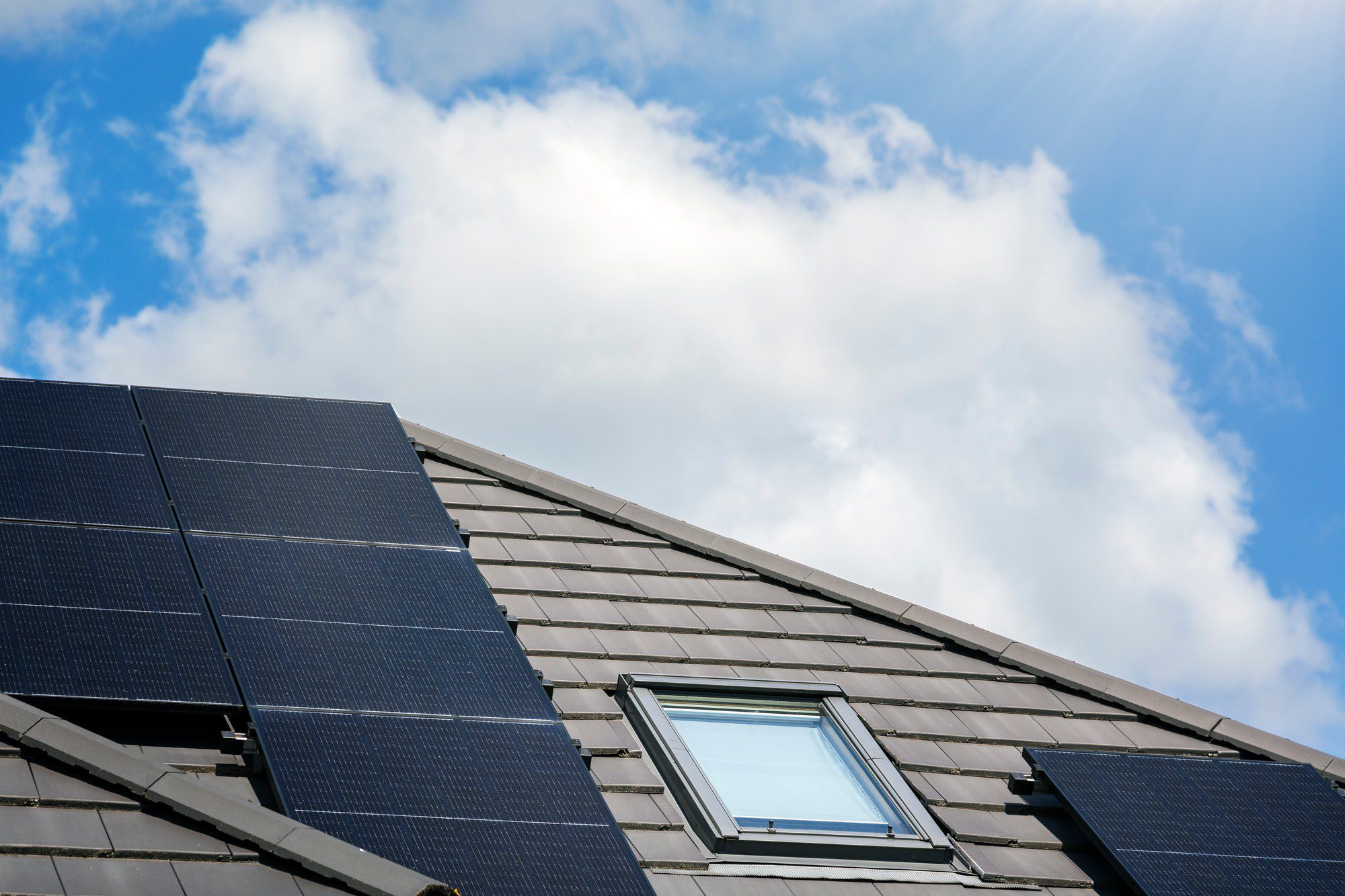 The image depicts a modern, residential house with a dark grey, sloped roof that is covered with multiple photovoltaic solar panels. The solar panels are rectangular and uniformly arranged, spanning nearly the entire surface of the roof. The house is set against a bright blue sky with minimal clouds, suggesting a sunny day. In the background, along the perimeter of the house, there is lush, green foliage indicating a well-maintained garden or surrounding area. The structure of the house is contemporary, with clean lines and a focus on sustainability as evidenced by the solar panel installation.