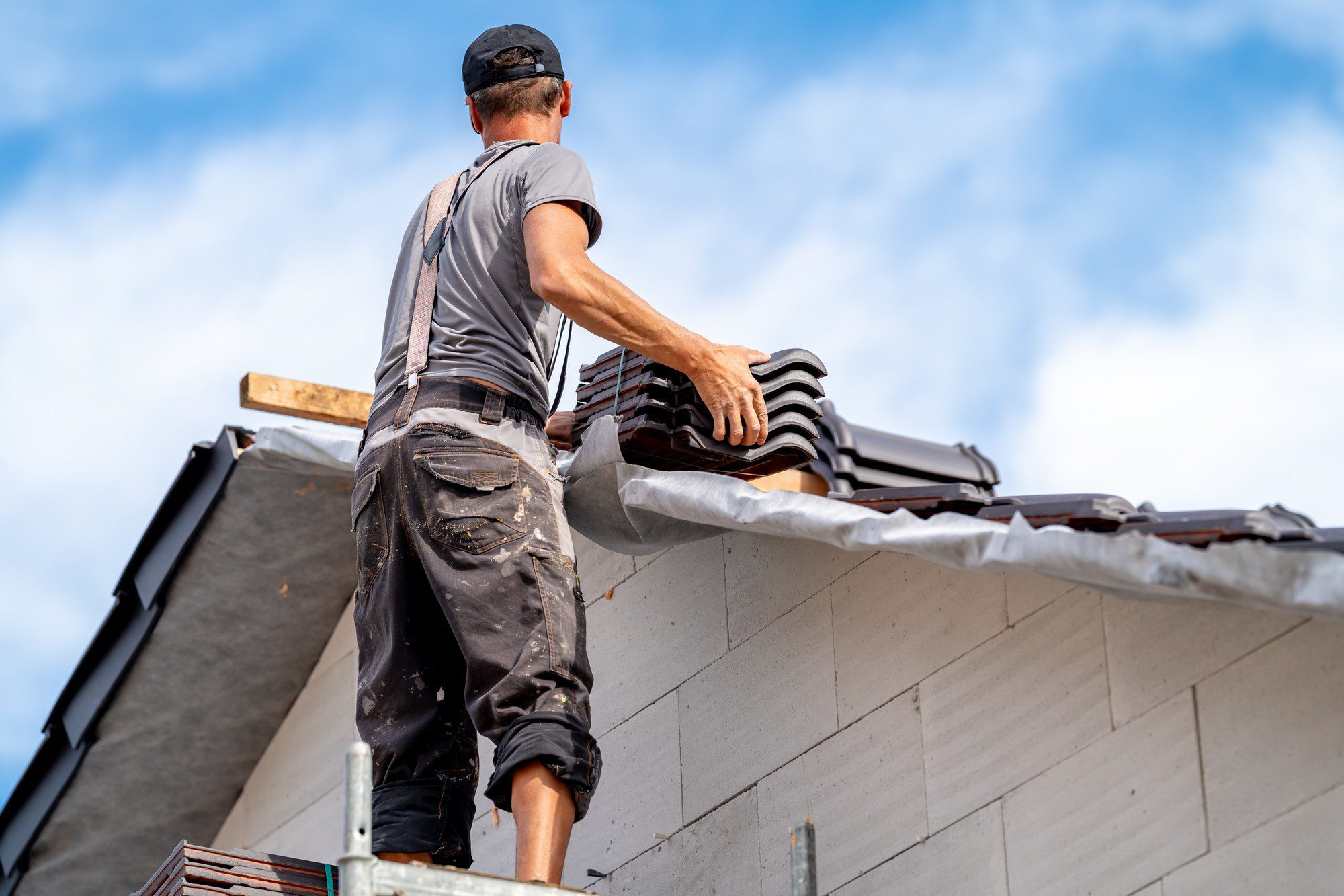 The image depicts a professional roofer working diligently atop the roof of a residential family house. The scene is set on a clear day with ample daylight, suggesting an optimal working environment. The roofer, dressed in appropriate safety gear including a high-visibility vest, helmet, gloves, and sturdy work boots, is focused on his task. He is in the process of laying down or securing roofing materials, which could be shingles, tiles, or membranes. The house appears to be a medium-sized, suburban home with a pitched roof. Surrounding the house are leafy green trees and a manicured lawn, contributing to the picturesque residential neighborhood setting. Various tools such as a hammer, a toolbox, and materials are scattered neatly on the roof around the roofer, indicating meticulous and organized work. The bright weather highlights the worker’s concentration and dedication to his craft, embodying a snapshot of routine but essential home maintenance.